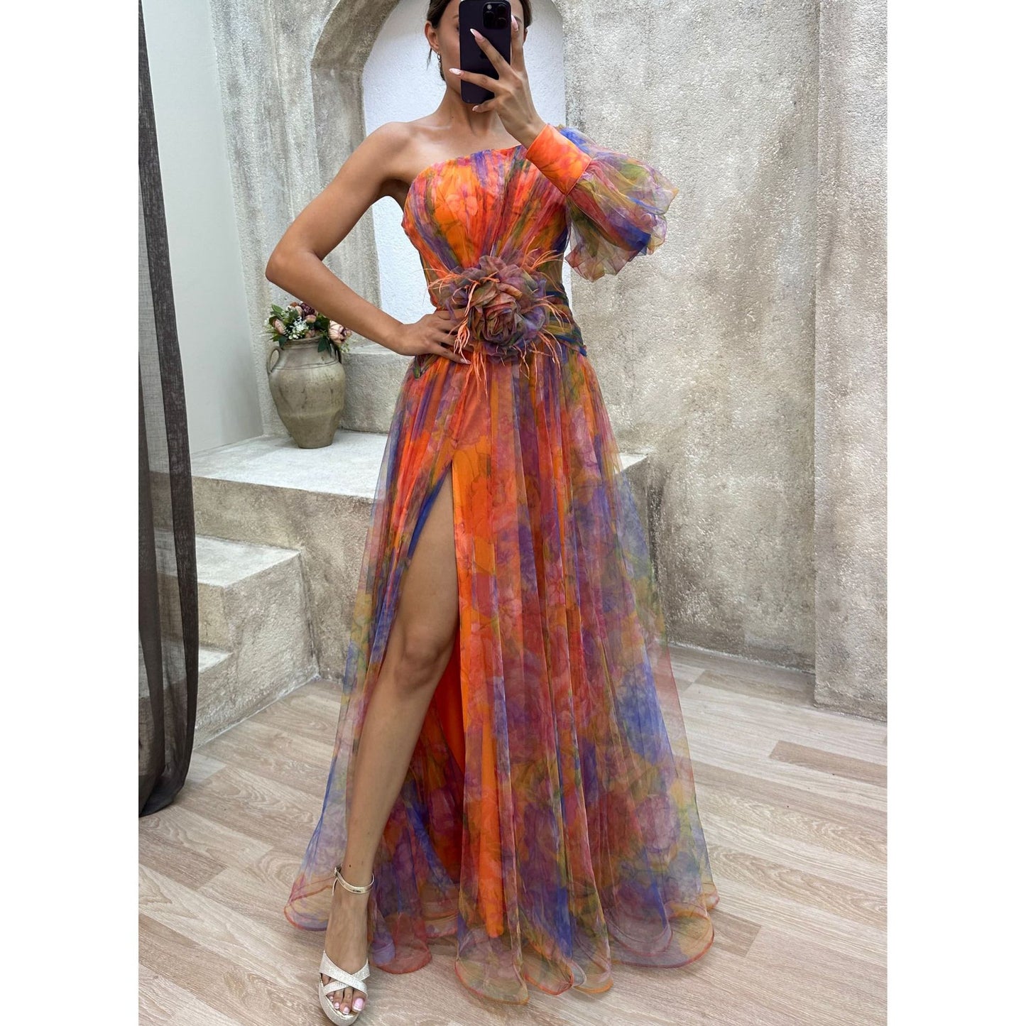 Woman wearing a colorful one-shoulder dress with a high slit, standing in a room with a mirror and decorative items.