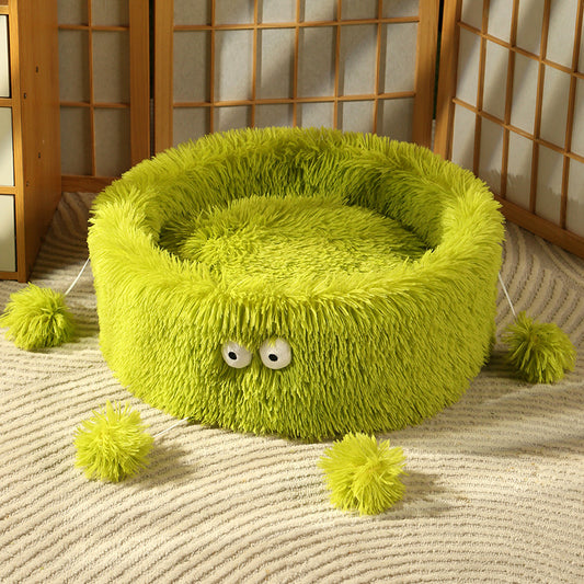 Green fluffy pet bed with eyes and pom-poms on a textured floor.