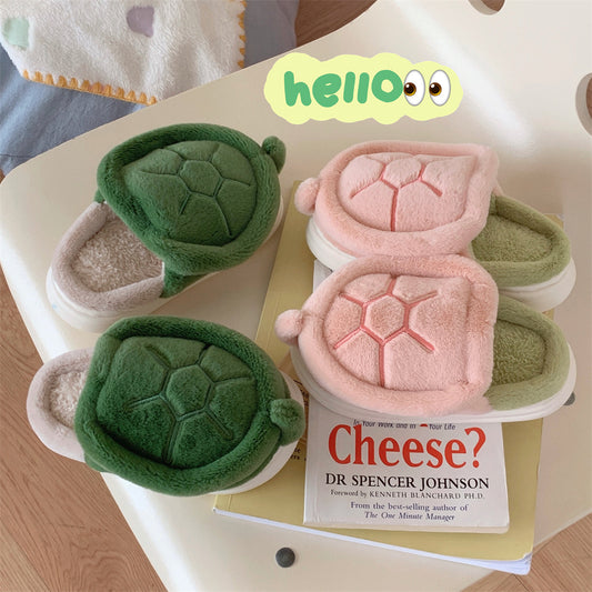 Cute plush turtle slippers in green and pink with soft inner lining, placed on a table with a children's book